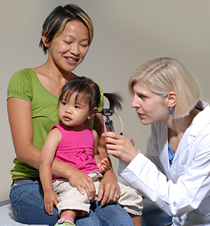 Una niña pequeña sentada en la falda de una mujer mientras la médica le examina el oído con un otoscopio.