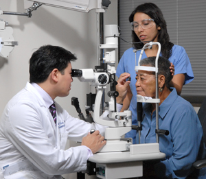 Oftalmólogo examinando el ojo de una mujer con lentes y una lámpara de hendidura. Otro proveedor de atención médica sostiene la cabeza de la paciente para que no se mueva.