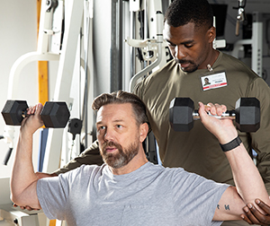 Un hombre entrenando a otro hombre que hace ejercicios de fortalecimiento muscular con mancuernas.