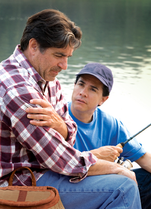 Hombre sentado junto a un lago con una caña de pescar; se está tomando de un brazo y se lo ve angustiado. Un hombre más joven está sentado junto a él y se lo ve preocupado.